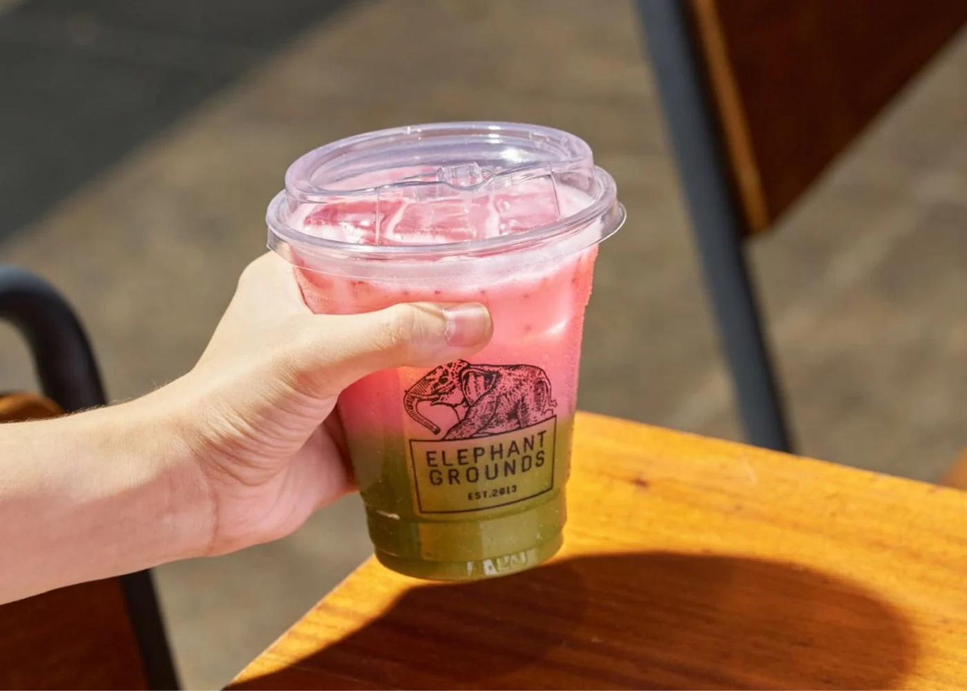 A hand holding a pink and green layered drink in an Elephant Grounds cup.